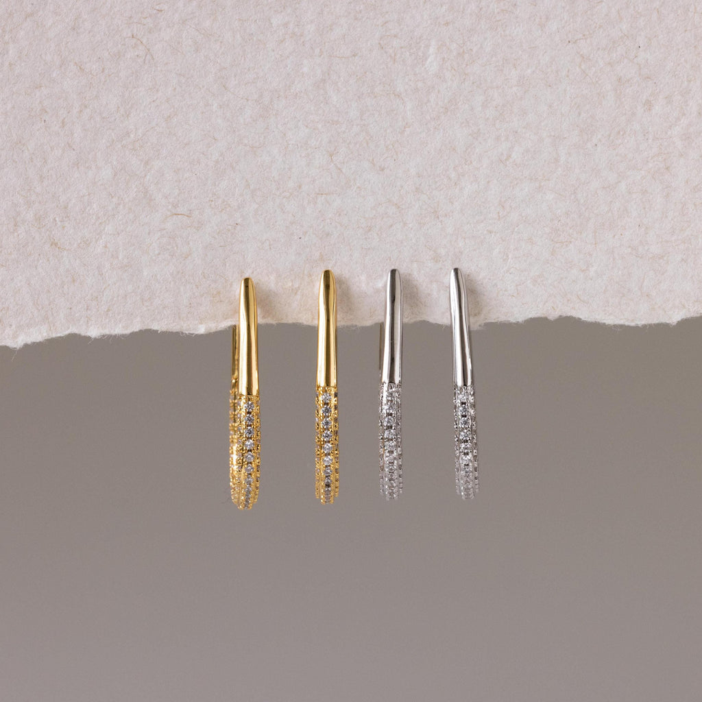 The Bold Pave Oval Hoops set features two pairs of thin, textured statement earrings in gold and silver, displayed on a torn paper background—ideal for adding a touch of sparkle to any outfit.