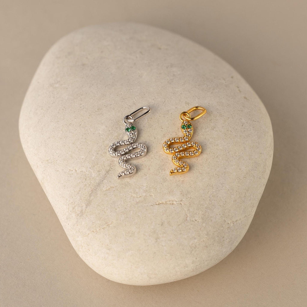 One silver and one diamond snake charm pendant lay side by side. The design is a twisting snake with diamond gemstones encased on its body and emerald gemstones for its eyes