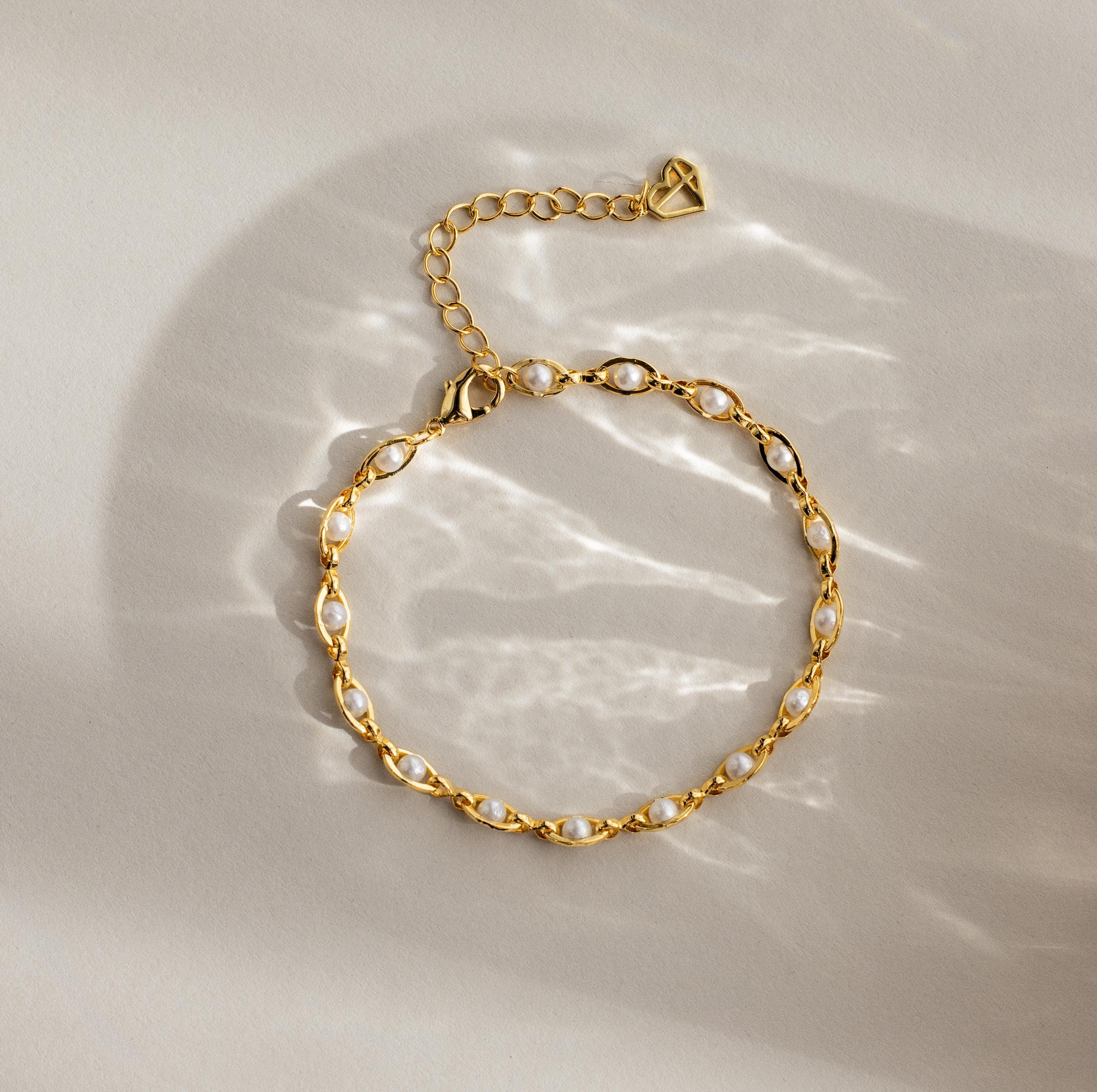 Eternity Pearl Bracelet: This elegant piece showcases pearls on a gold infinity chain, beautifully displayed flat on a beige surface with gentle, soft light reflections.