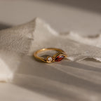 The Toi et Moi Infinity Birthstone Ring features a red marquise gemstone and a round clear stone, displayed on textured cream fabric—a perfect token of love.
