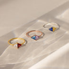 The Toi et Moi Triangle Birthstone Ring features colorful geometric gemstones set in striking gold, rose gold, and silver bands, displayed on a beige surface.