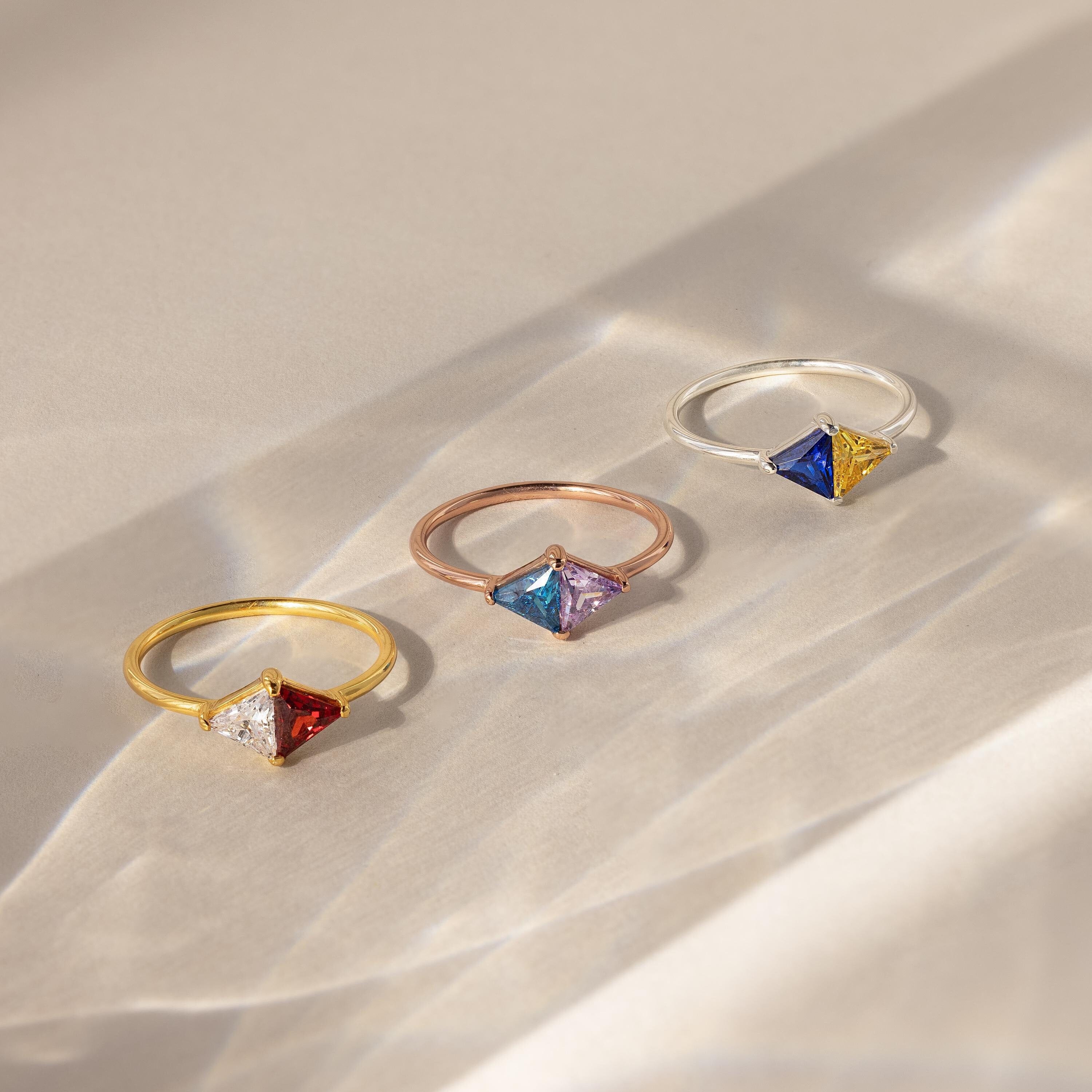 The Toi et Moi Triangle Birthstone Ring features colorful geometric gemstones set in striking gold, rose gold, and silver bands, displayed on a beige surface.