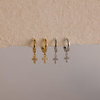 Two gold and two silver Pave Cross Huggies, each with a delicate dangling cross pendant, are beautifully displayed on a textured surface.