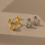 Two pairs of Medium Diamond Bezel Flatback Studs, one gold and one silver, with round clear stones rest on a beige surface—elegant additions to your jewelry collection.