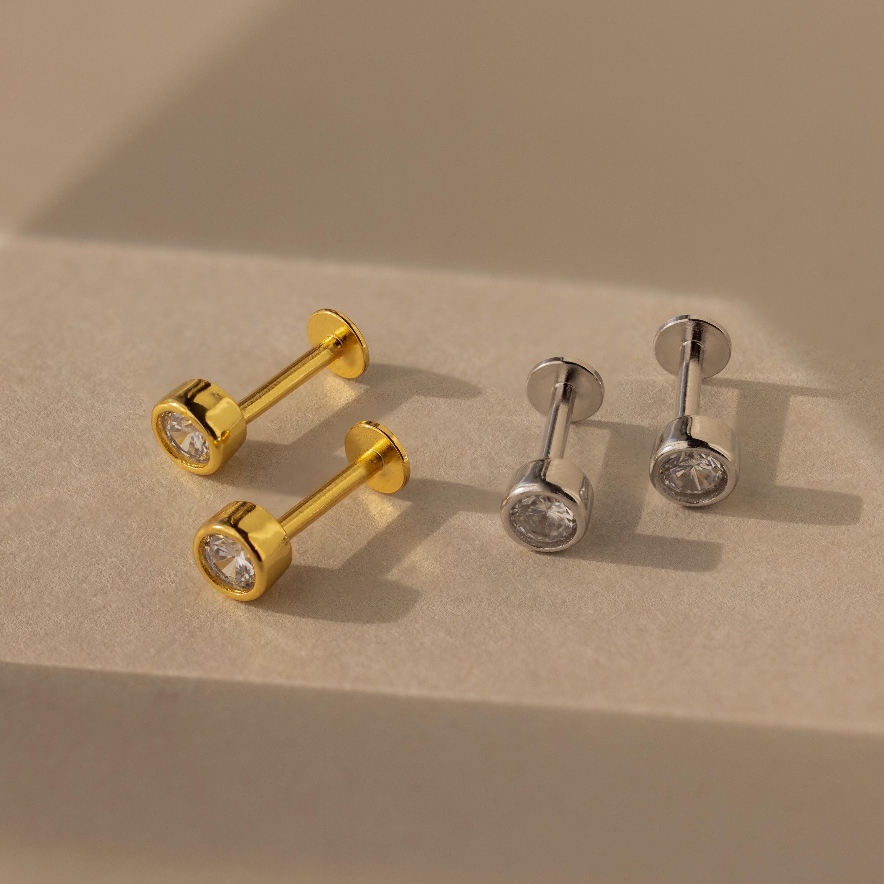 Two pairs of Medium Diamond Bezel Flatback Studs, one gold and one silver, with round clear stones rest on a beige surface—elegant additions to your jewelry collection.