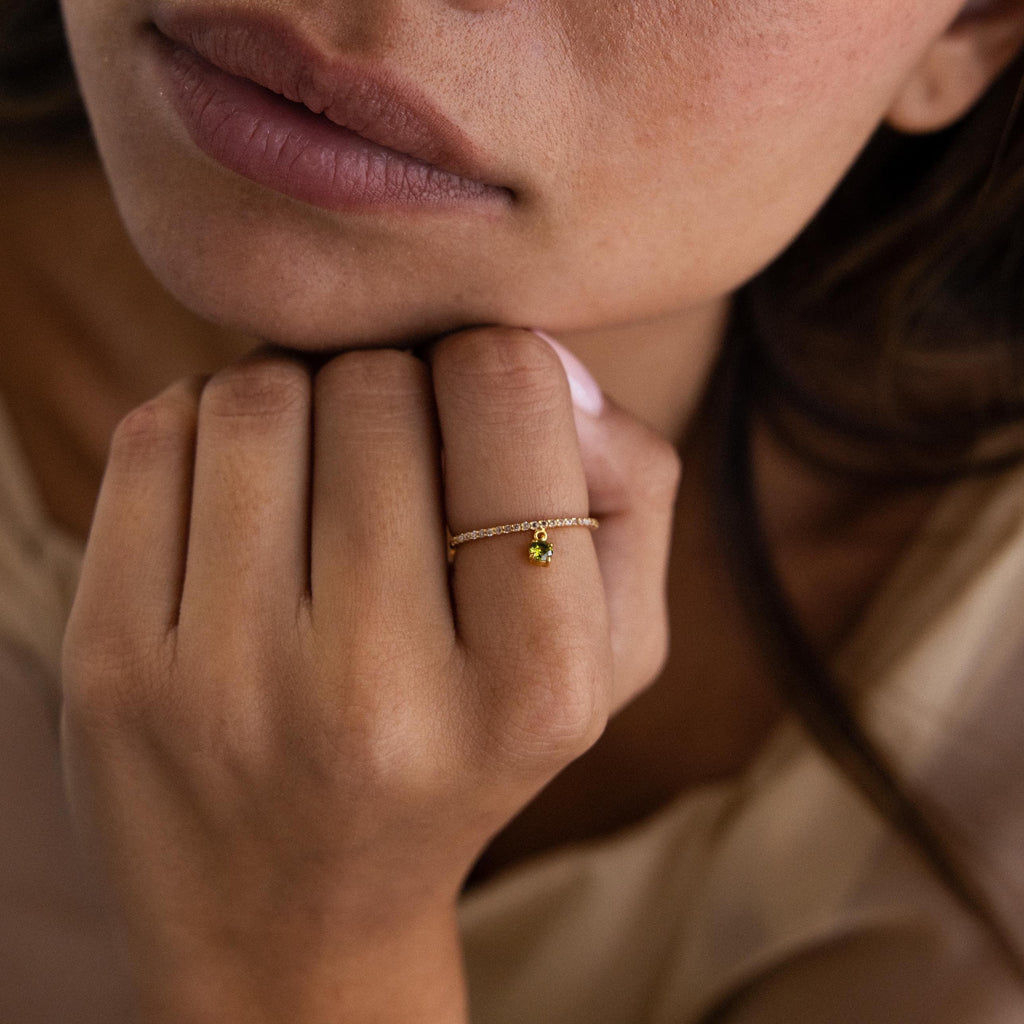 A woman rests her chin on her hand, wearing the Pave Birthstone Drop Ring featuring a delicate gold band and a small green gemstone.