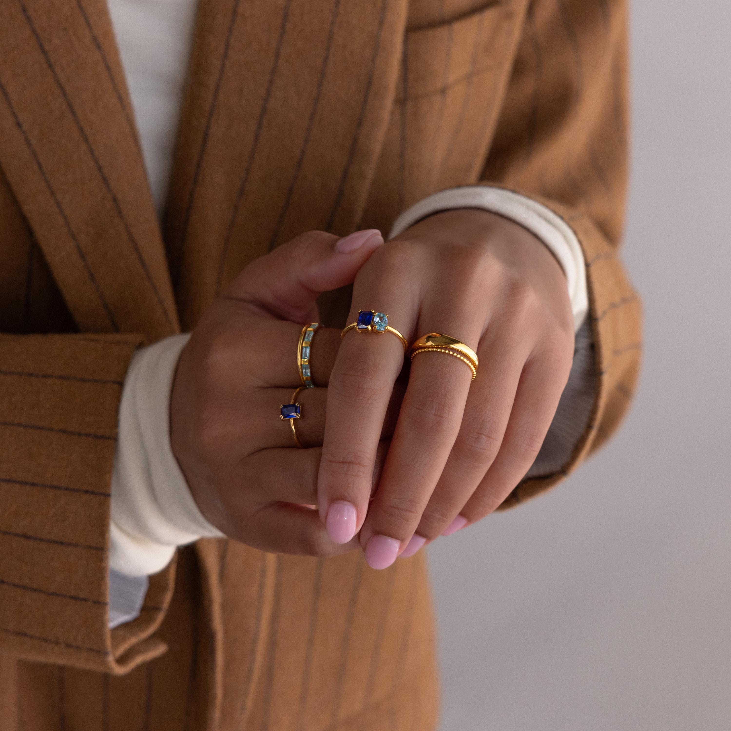 Hands with pink nails showcase multiple gold rings, featuring the Toi et Moi Oval Birthstone Ring, paired with a brown pinstripe blazer and white top.