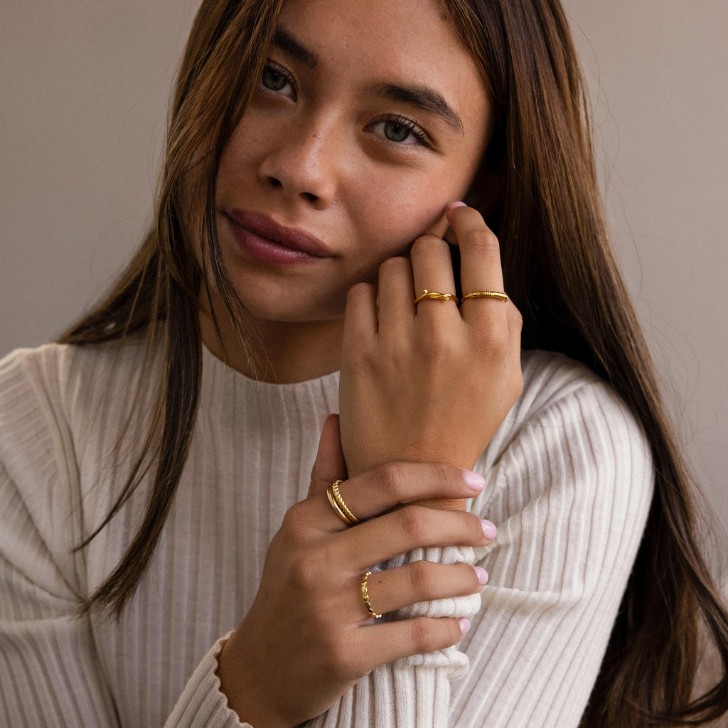 A woman in a white ribbed top showcases gold personalized jewelry, featuring the Dainty Morse Code Ring on her finger, as she looks at the camera with a slight smile.