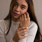A woman in a white ribbed top showcases gold personalized jewelry, featuring the Dainty Morse Code Ring on her finger, as she looks at the camera with a slight smile.