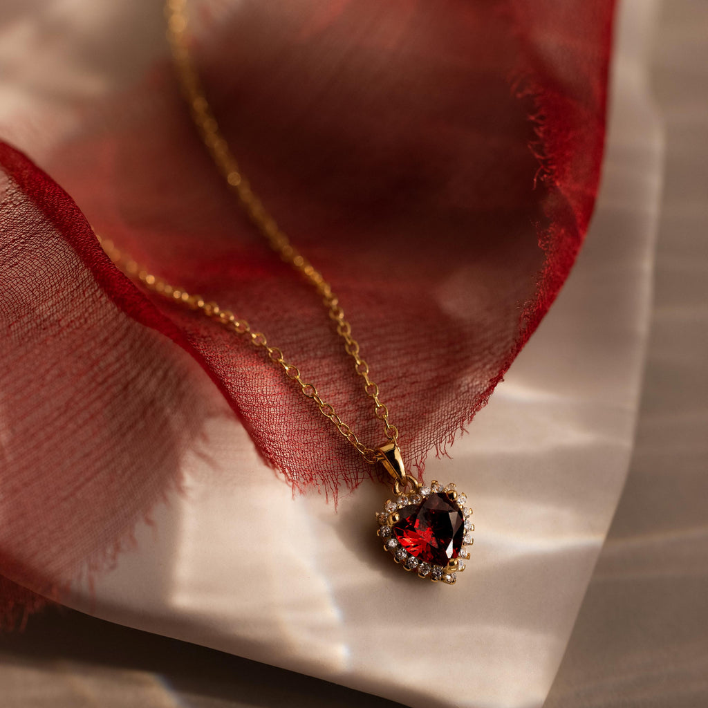 The Pave Garnet Heart Necklace features a heart-shaped red gemstone pendant, beautifully displayed on soft red fabric and a marble surface.