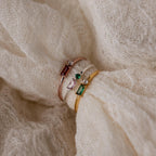 The Toi et Moi Baguette Birthstone Ring features sparkling colored gemstones and diamonds, elegantly displayed on textured cream fabric.