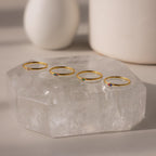 Four delicate gold rings, featuring the Mini Toi et Moi Birthstone Ring, display colorful gemstones and sparkle on a large clear crystal slab.