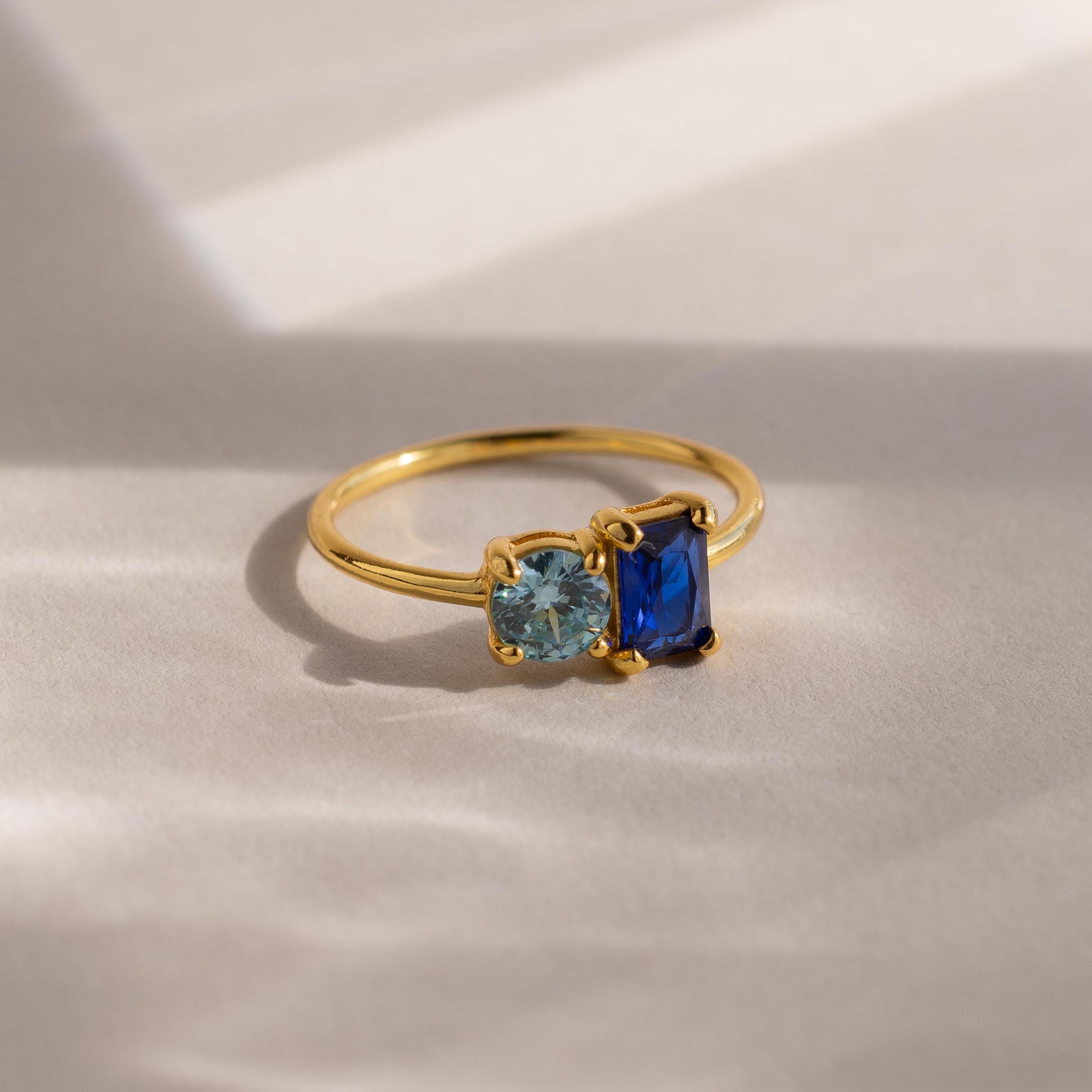 The Toi et Moi Emerald Birthstone Ring features a gold band with one light blue round gem and one dark blue rectangular gem, elegantly displayed on a light surface.