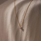 Baguette Birthstone Drop Necklace with a small purple gemstone pendant, shown on a textured beige surface.