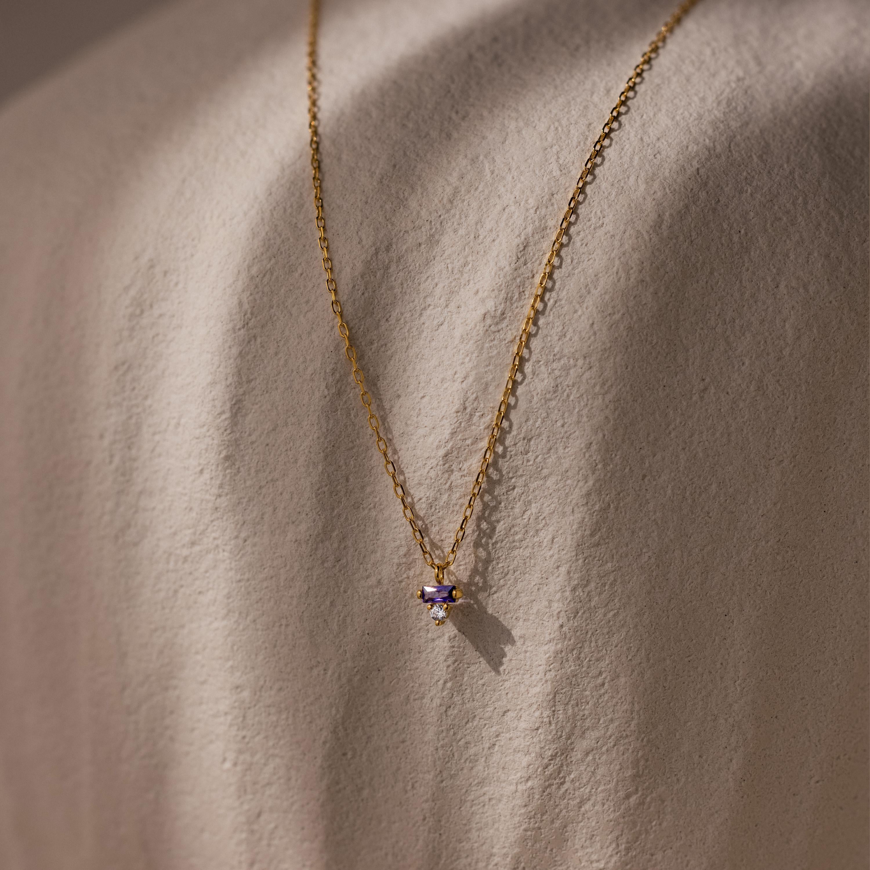 Baguette Birthstone Drop Necklace with a small purple gemstone pendant, shown on a textured beige surface.