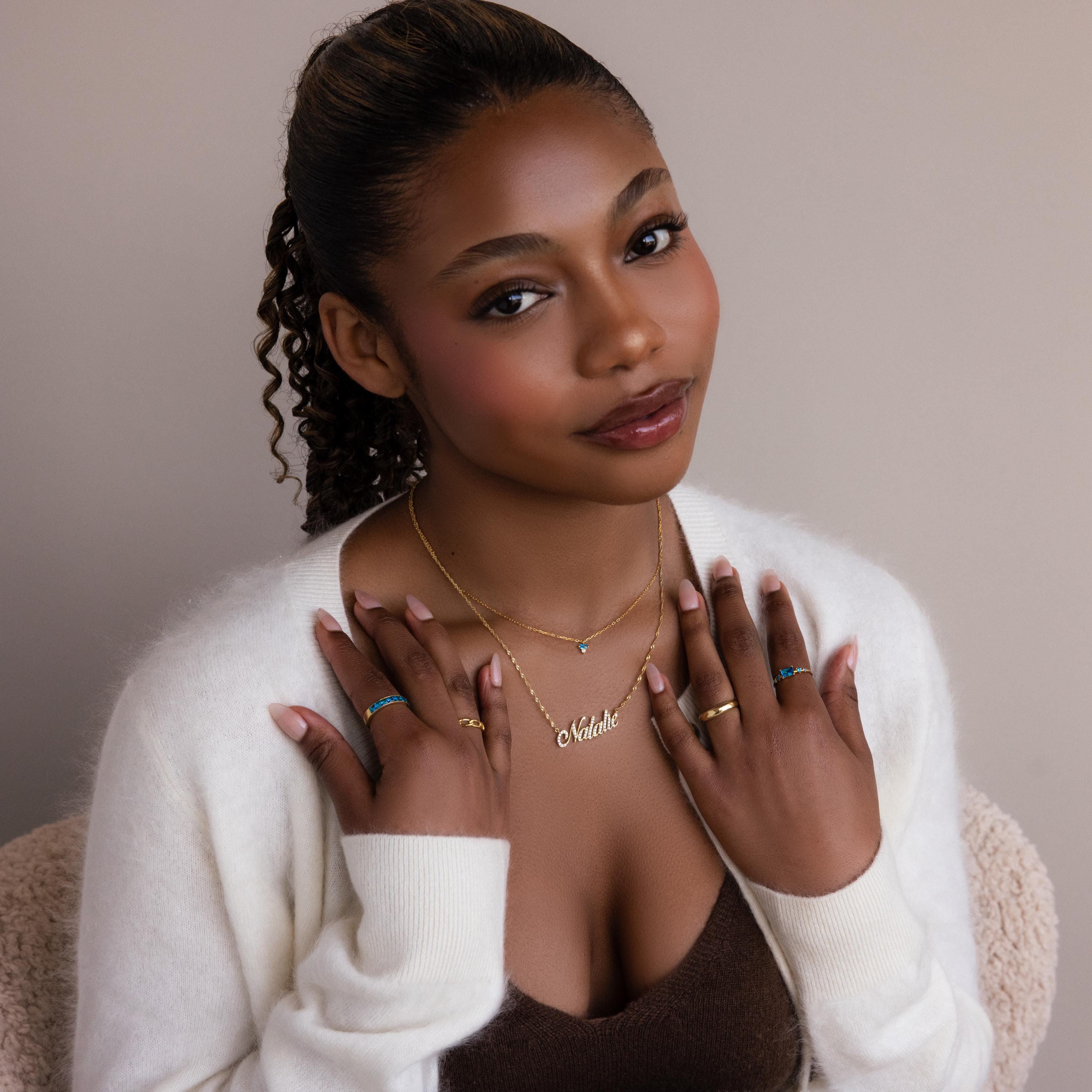 Woman with curly hair, wearing a Baguette Birthstone Drop Necklace and a white cardigan, poses with hands near her chest and smiles softly.