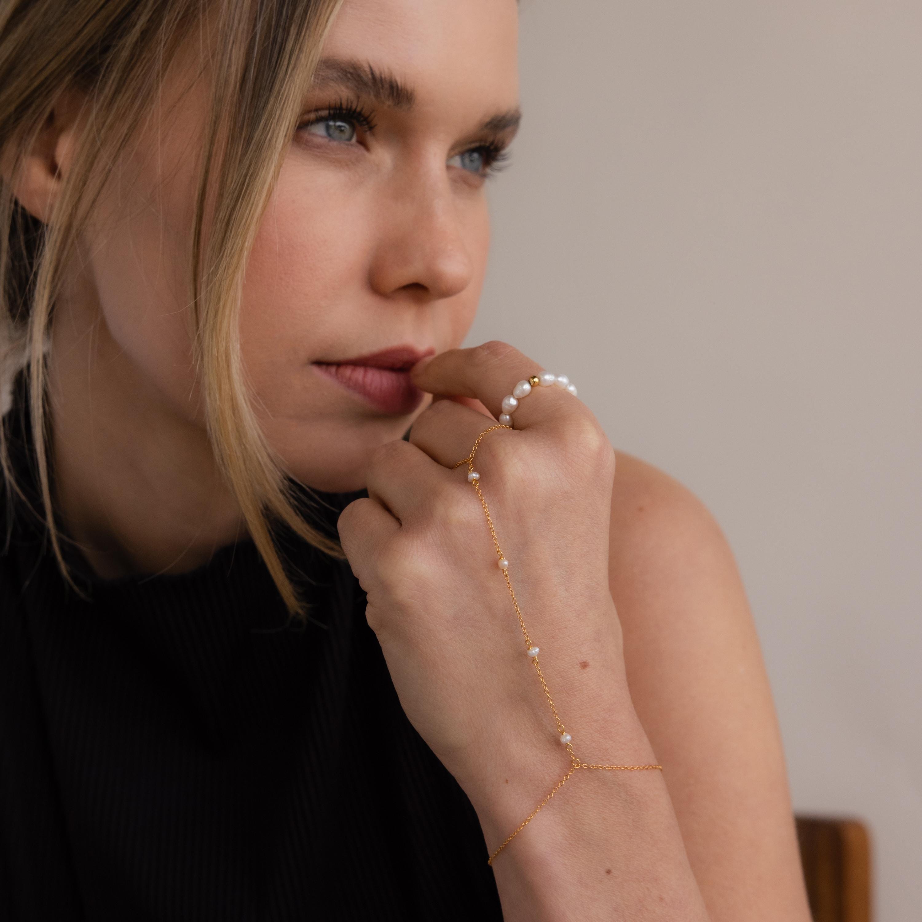 A blonde woman wears the Pearl Hand Chain Bracelet, featuring gold and pearls, as she gazes thoughtfully to the side—embodying effortless summer elegance.