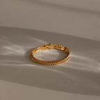 The V-Link Chain Tennis Bracelet, featuring gold sculptural links in a herringbone design, is displayed on a beige surface under soft natural light with gentle shadows.
