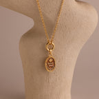 The Love Poem Pendant Necklace features engraved details and is showcased on a beige textured stand with a neutral background—a timeless love token.