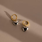 The Mixed Metal Heart Huggies feature gold hoops with silver puff heart charms, displayed on a beige surface with soft shadows to highlight their elegant, contemporary style.