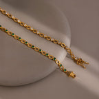 The Emerald Chain Tennis Bracelet featuring emerald stones, along with a white stone chain bracelet, are displayed on a round beige surface.