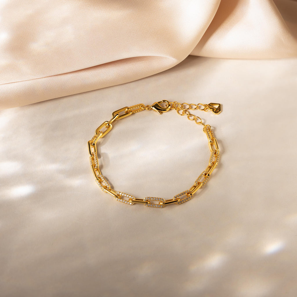 The Pave Paperclip Chain Bracelet features rectangular links with small crystals, elegantly displayed on a soft beige fabric background.