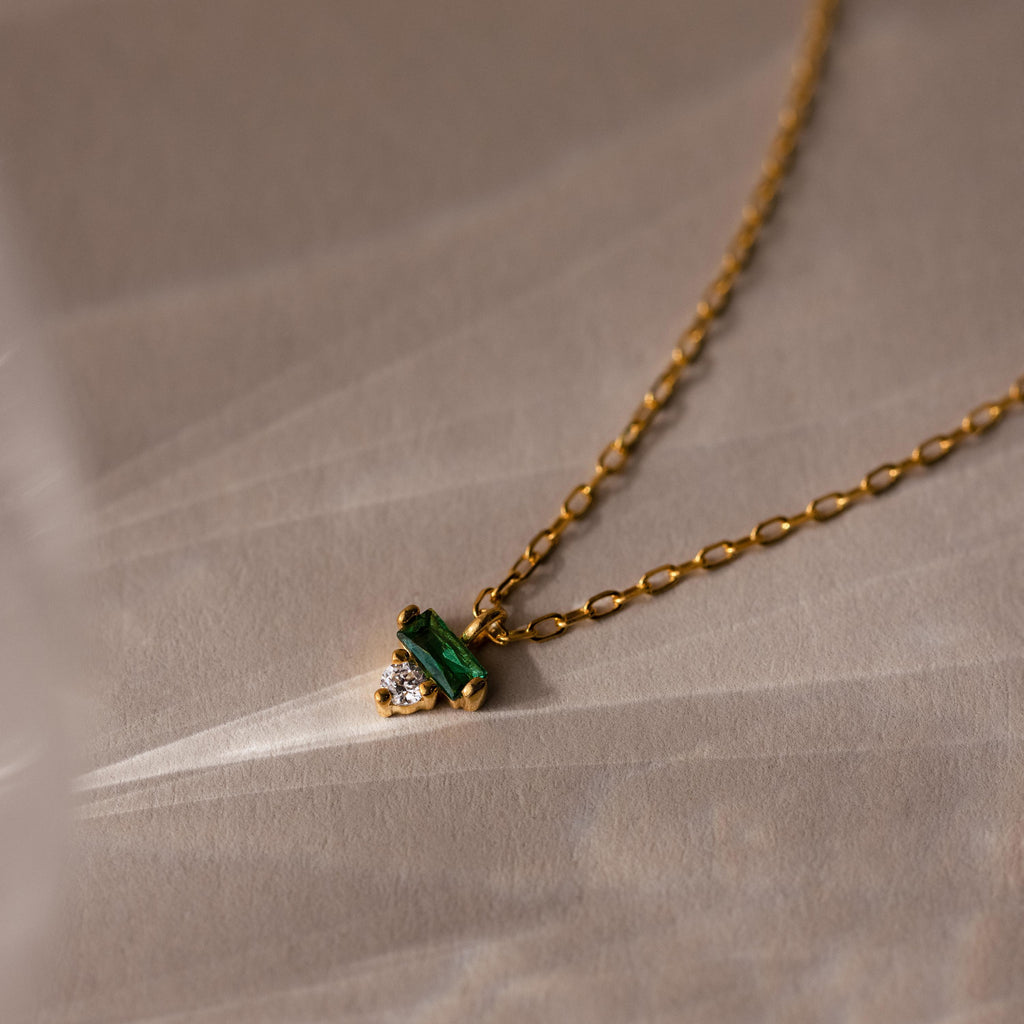 The Baguette Birthstone Drop Necklace features a gold chain with a small green and clear gemstone pendant, displayed on a beige textured background.