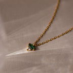 The Baguette Birthstone Drop Necklace features a gold chain with a small green and clear gemstone pendant, displayed on a beige textured background.