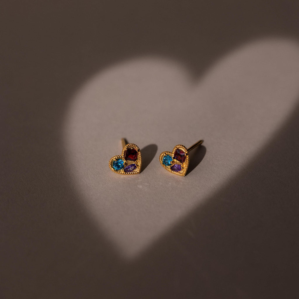 Two gold heart-shaped earrings lit by a heart-shaped light on a gray surface. Discover the Mixed Birthstone Heart Studs—customizable with your choice of gemstones for a unique touch.