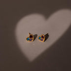 Two gold heart-shaped earrings lit by a heart-shaped light on a gray surface. Discover the Mixed Birthstone Heart Studs—customizable with your choice of gemstones for a unique touch.