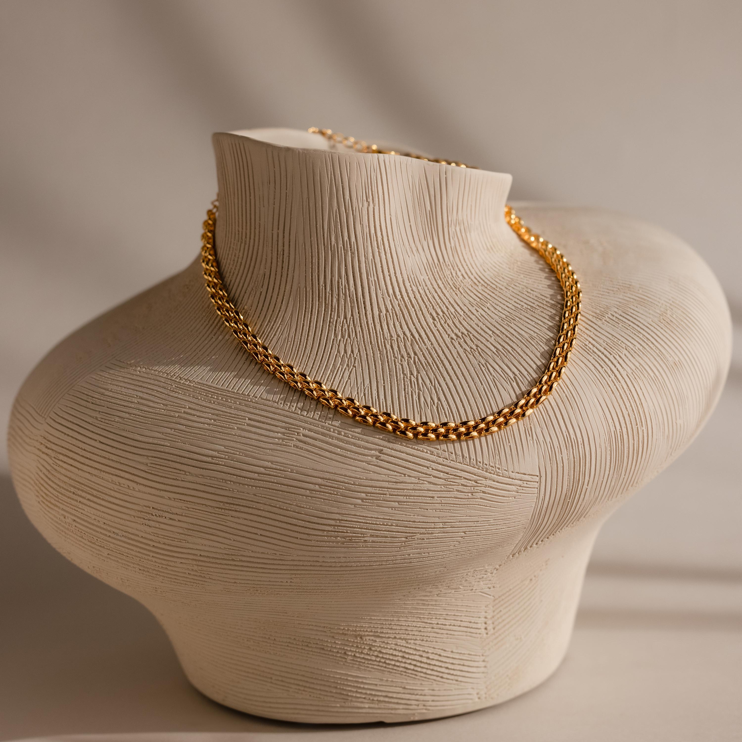 The Strap Necklace, a luxury gold chain with interwoven links, is displayed on a textured beige sculpted bust against a neutral background.