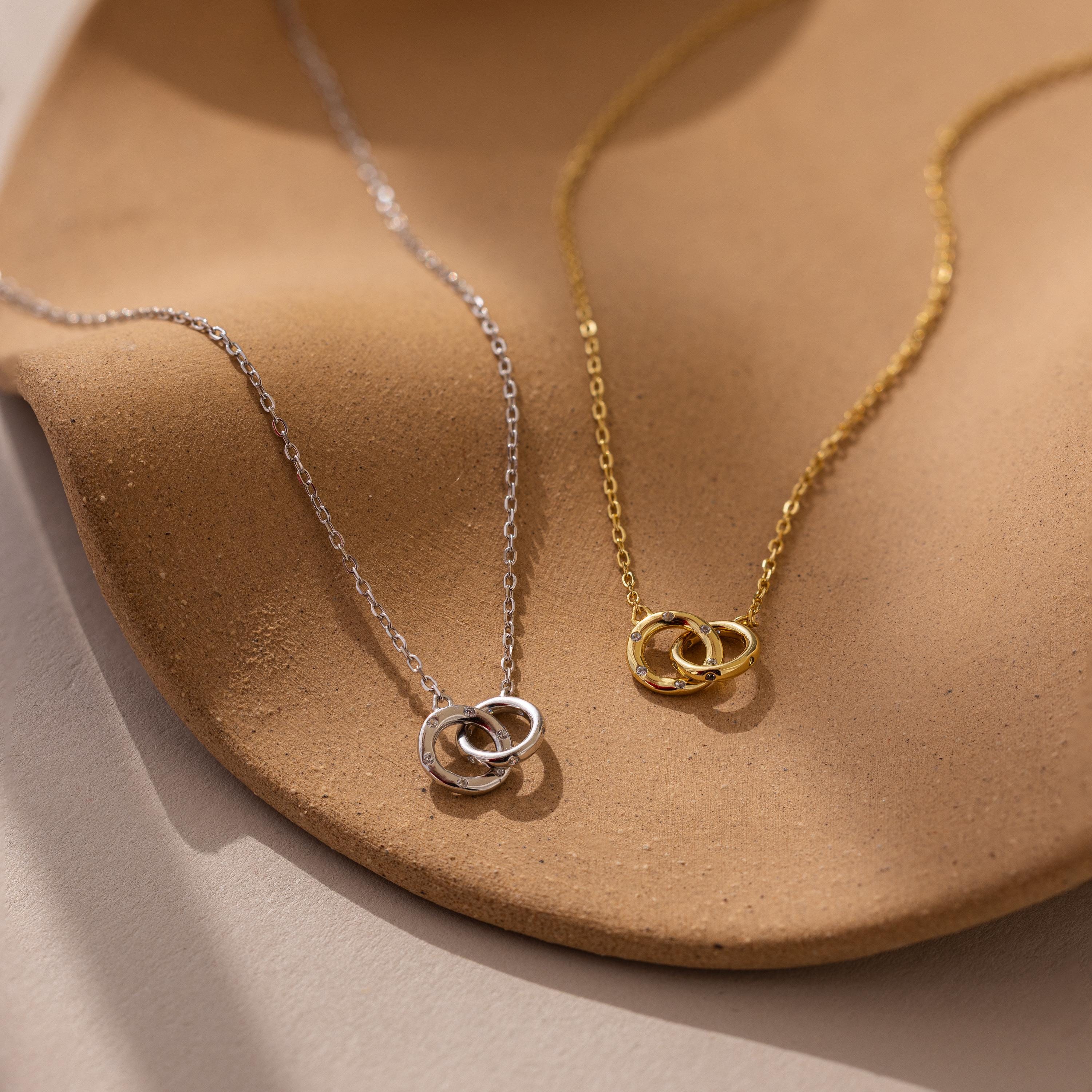The Pave Interlocking Circle Necklace features one silver and one gold interlocking circle, symbolizing love and unity. Displayed on a textured beige surface, each necklace sparkles with delicate CZ diamonds.