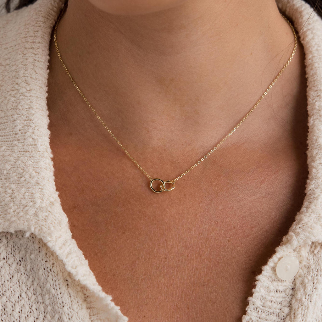 Wearing the Pave Interlocking Circle Necklace, featuring two linked gold rings symbolizing love, over a cream textured shirt.