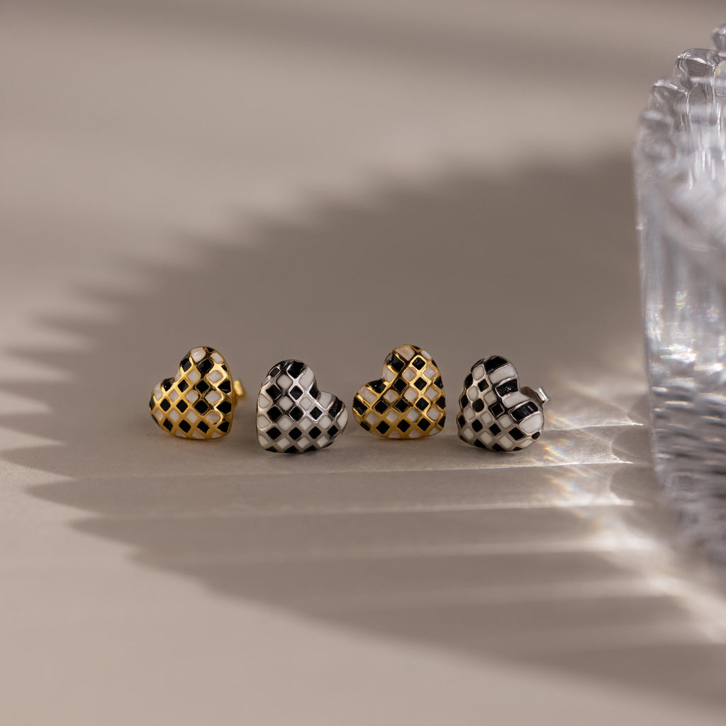 A set of four Checkered Heart Studs featuring retro-inspired gold and silver patterns is displayed beside a textured glass, with shadows cast across the surface.