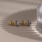 A set of four Checkered Heart Studs featuring retro-inspired gold and silver patterns is displayed beside a textured glass, with shadows cast across the surface.
