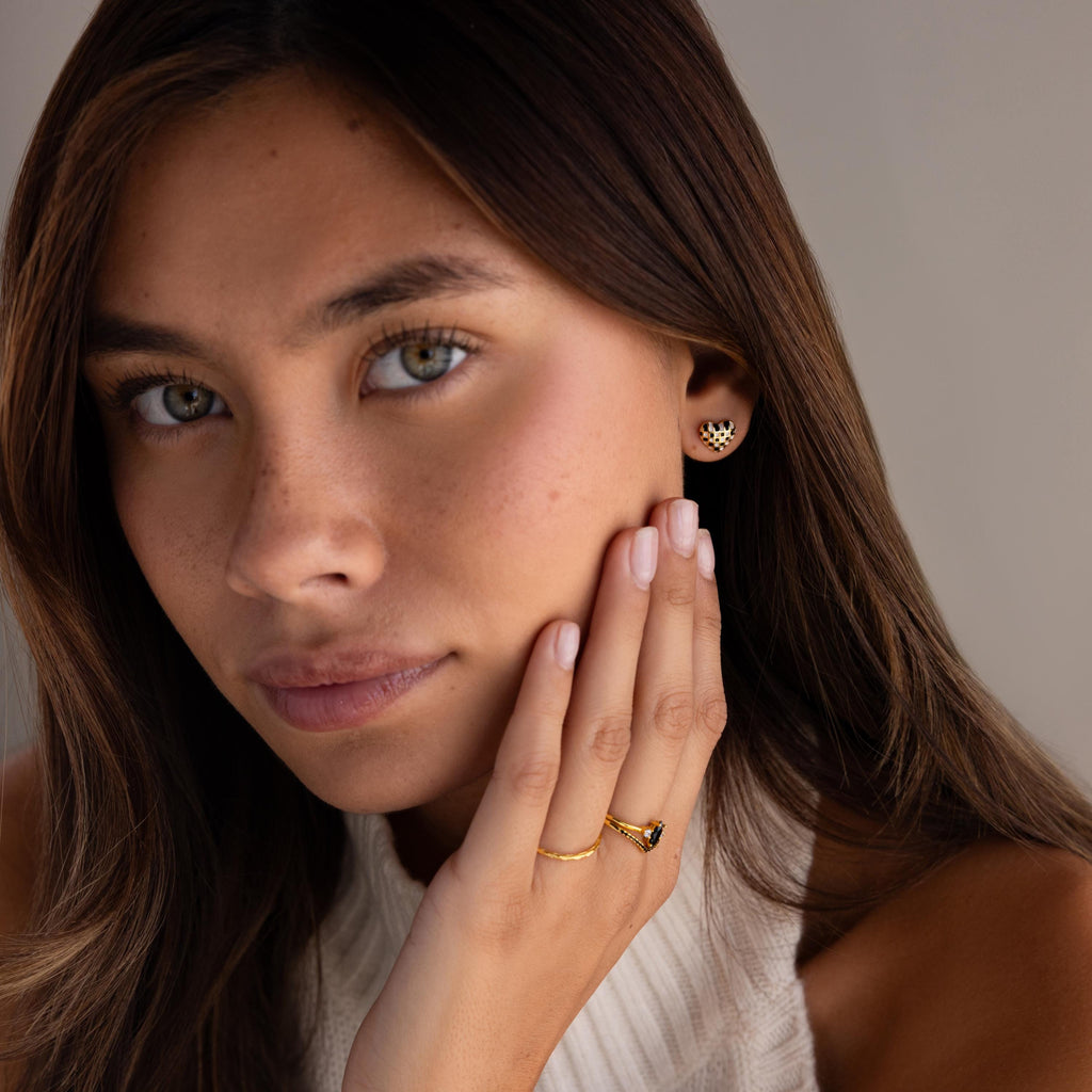 A woman with light brown hair wears gold jewelry, including the Checkered Heart Studs, a ring, and a sleeveless white top.