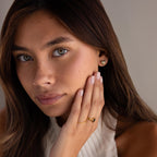 A woman with light brown hair wears gold jewelry, including the Checkered Heart Studs, a ring, and a sleeveless white top.