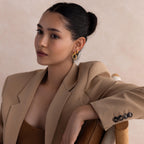 A woman with dark hair in a bun, wearing a tan blazer and Bold Duo Knot Earrings, sits elegantly on a chair.