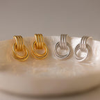 The Ribbed Duo Knot Earrings, in gold and silver, are statement pieces that bring a modern touch to any outfit. Shown on a round iridescent dish, these stylish earrings elevate your look.