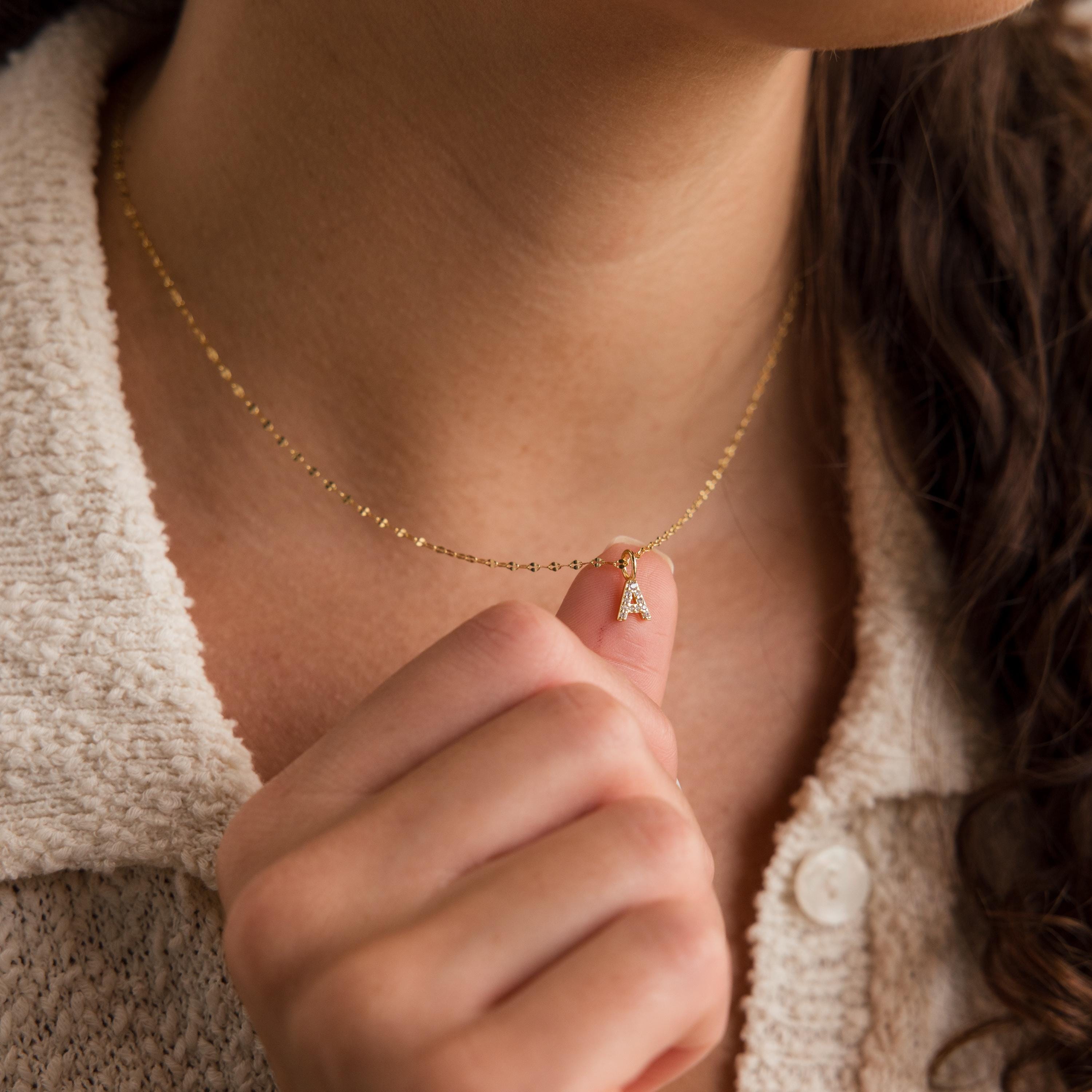 Gold initial pendant necklace with a pavé crystal letter "A" charm, worn on a delicate beaded chain and held between fingers.