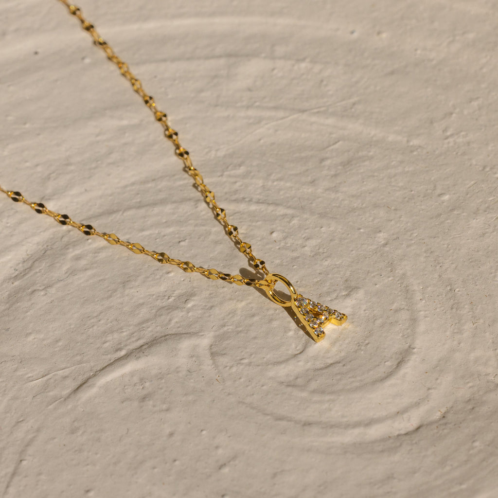 The Dainty Pave Letter Necklace in 18K Gold features a sparkling CZ pavé letter pendant, elegantly displayed on a textured beige surface.