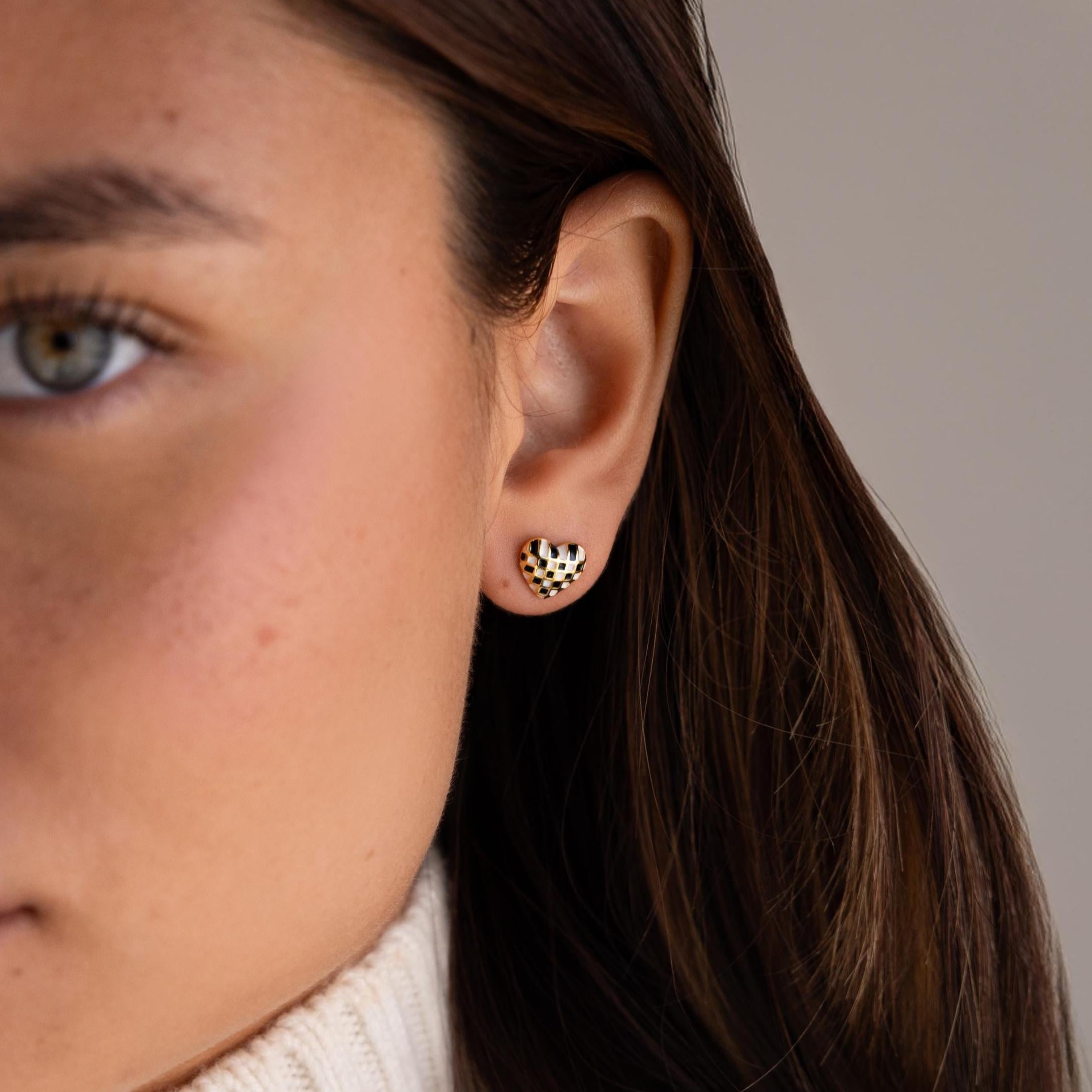 Close-up of a woman's ear wearing Checkered Heart Studs—a small, gold earring with a checkered pattern—accented by brown hair and a beige sweater.