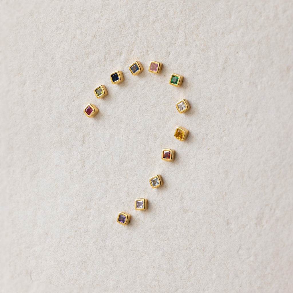 Square Bezel Pink Tourmaline Flatback Studs, along with other small colorful gemstones, are arranged in a question mark shape on a textured beige surface.