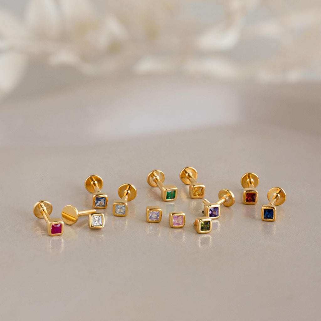 Nine pairs of Square Bezel Birthstone Flatback Studs, featuring gold settings with colorful gemstones, are displayed on a smooth neutral surface.