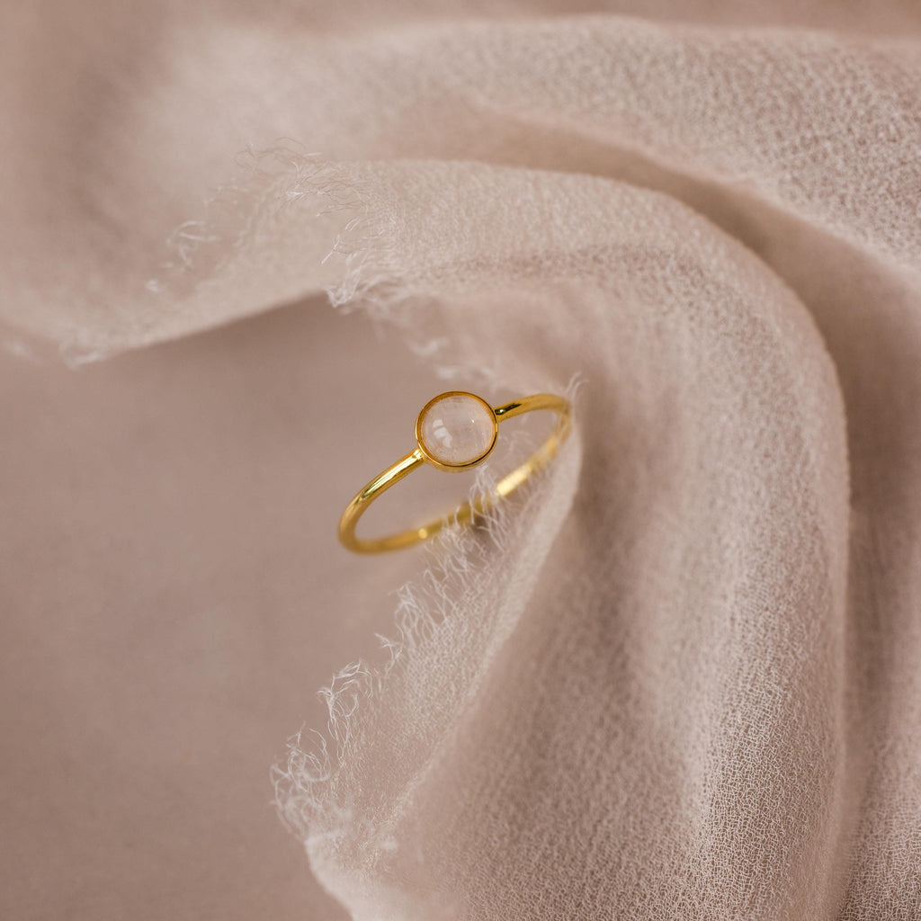 The Dainty Round Moonstone Ring features a subtle round gemstone, exuding understated elegance and grounding vibes when displayed on soft, textured fabric.