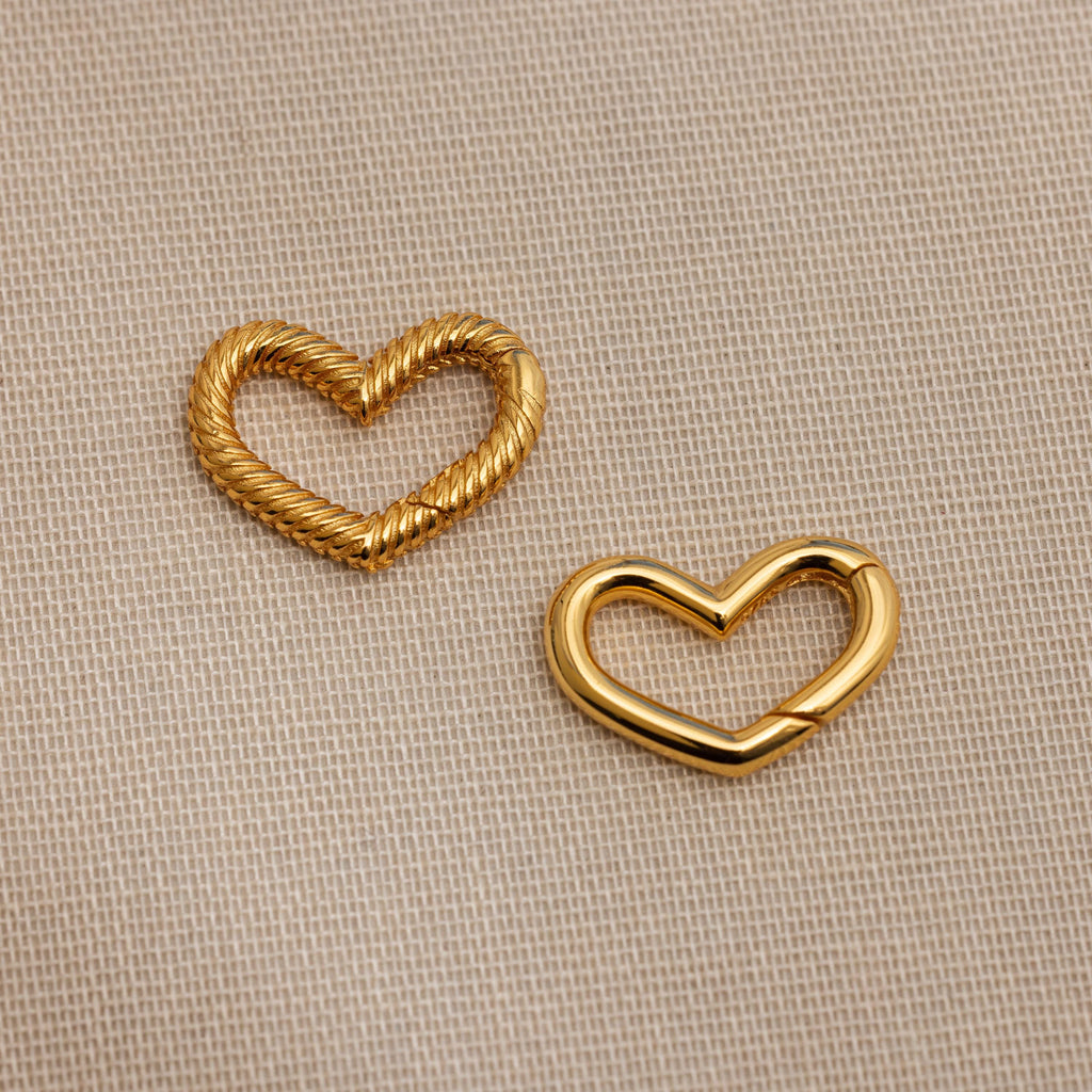 The Heart Carabiner Set in 18K Gold features two heart-shaped charms—one twisted, one smooth—perfect as a bracelet connector or jewelry set accent, displayed on beige textured fabric.
