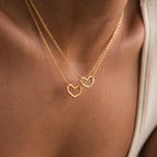 Two Heart Carabiner Set in 18K Gold necklaces rest on a person's neck, each with a delicate heart-shaped carabiner. The close-up captures the jewelry's charm and elegance against the skin, highlighting this stunning set.