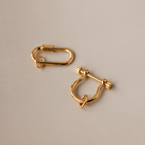 Two gold charm connectors—one oval and one U-shaped with a bar—rest on a smooth beige surface, perfect as necklace accessories or as part of the Bar & Carabiner Charm Set.