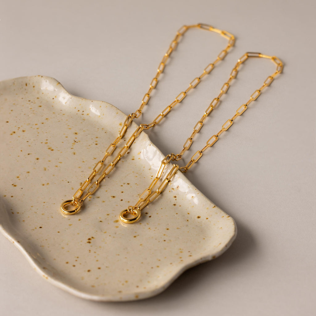 The Paperclip Clasp Necklace, a striking gold chain, is elegantly displayed on a speckled ceramic dish set against a beige background.