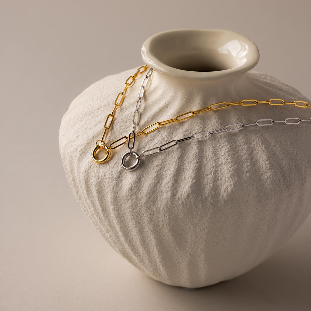 A white textured vase displays a Paperclip Clasp Necklace in bold gold and silver chain links, draped with collectible charms over its rim.